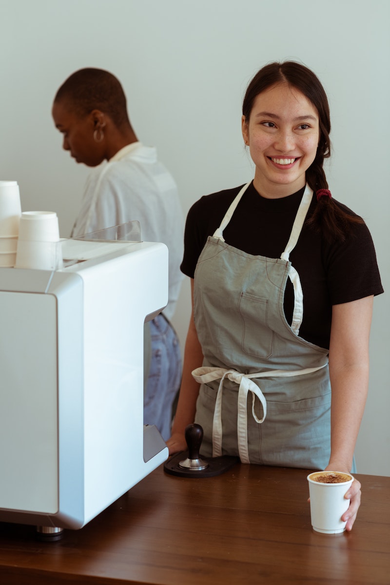 Positive Asian female coffee shop worker smiling and serving paper cup of freshly made coffee to customer with African American coworker behind coffeemaker in background
