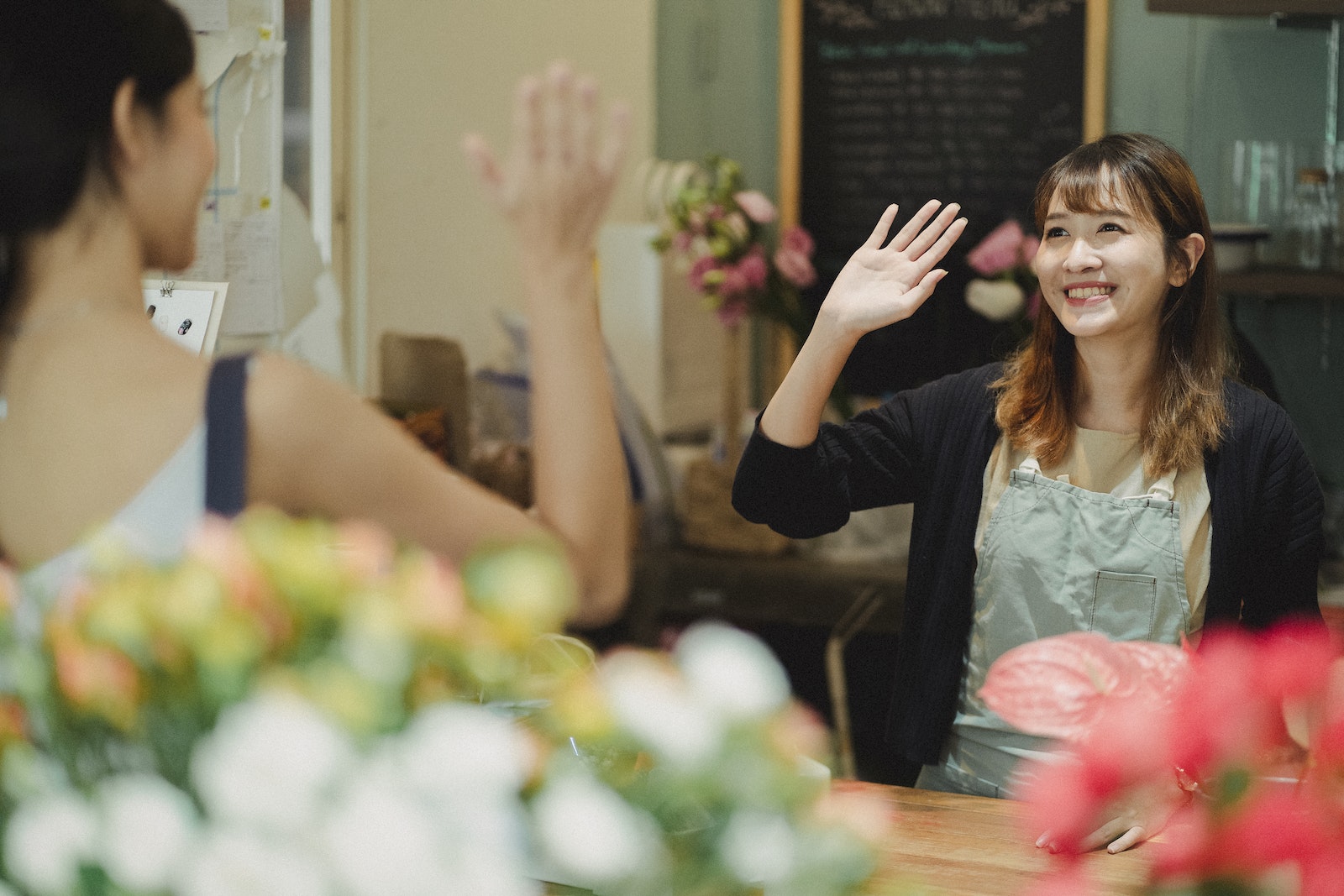 Positive Asian woman greeting client waving hand while working as florist in flower shop